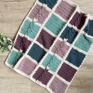 May include: A crocheted blanket with a patchwork design featuring squares in shades of teal, purple, and brown. The blanket is edged with a white border.