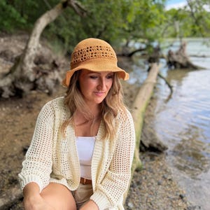 May include: A mustard yellow crocheted bucket hat with a decorative openwork band around the crown, paired with a cream-colored open-front cardigan featuring a loose mesh knit pattern, worn over a plain white top and light tan shorts.