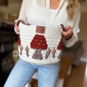 May include: A cream-coloured crochet basket featuring a pattern of dark red and brown mushrooms. The basket has a brown leather tag with the text "HAYLEY CROCHET" and brown leather side handles. The design is worked in a textured stitch pattern.