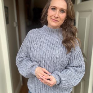 May include: A pale blue, chunky knit jumper with a high neck and long sleeves. The jumper is worn with blue jeans. The style is casual and comfortable, ideal for everyday wear. The sweater's texture is ribbed.