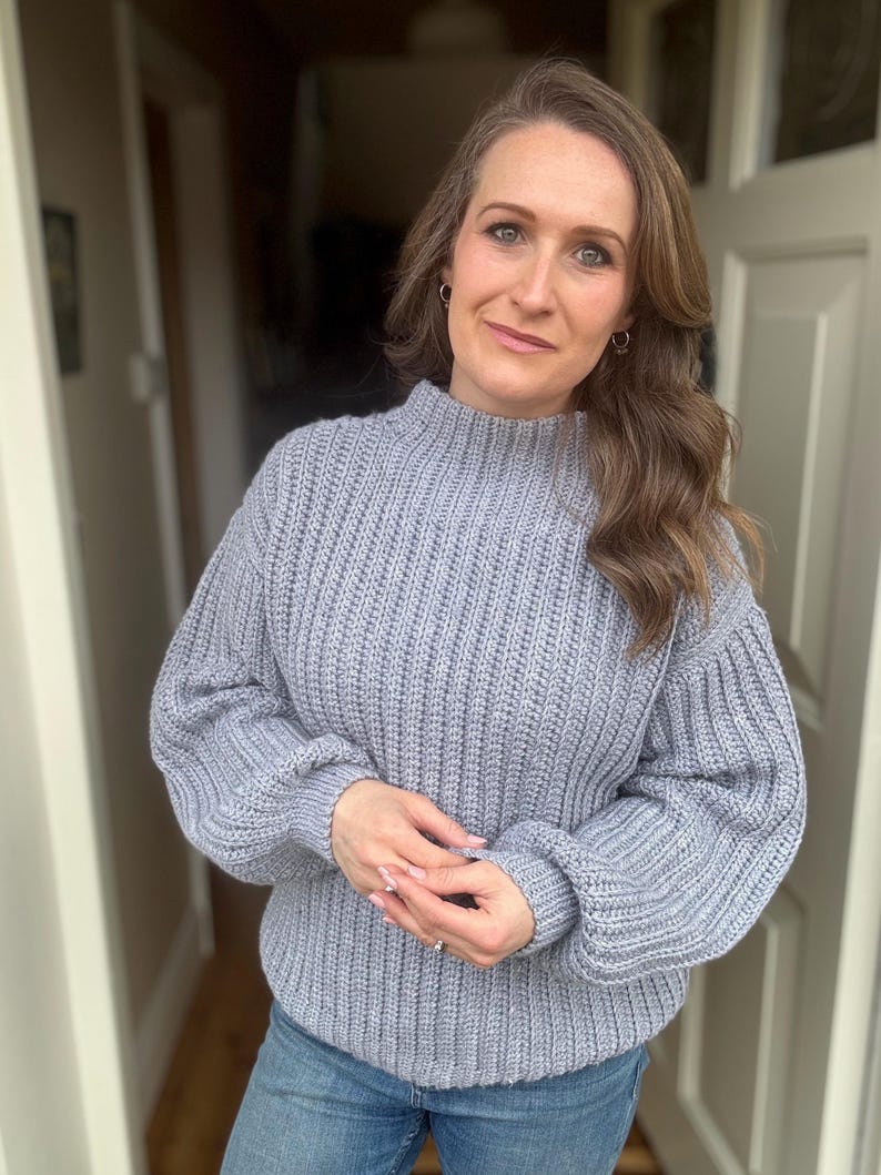 May include: A pale blue, chunky knit jumper with a high neck and long sleeves. The jumper is worn with blue jeans. The style is casual and comfortable, ideal for everyday wear. The sweater's texture is ribbed.