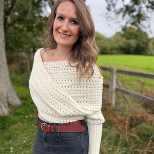May include: Cream-colored, off-the-shoulder crochet sweater with long sleeves. The sweater is paired with a brown leather belt and dark gray jeans. The background features a green field and a wooden fence.