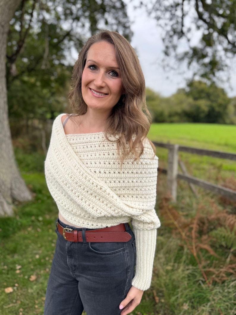 May include: Cream-colored, off-the-shoulder crochet sweater with long sleeves. The sweater is paired with a brown leather belt and dark gray jeans. The background features a green field and a wooden fence.