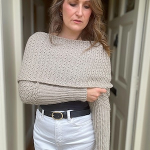 May include: A beige crochet sweater with long sleeves and a cowl neck. The sweater is worn over a black top and white jeans with a black belt.