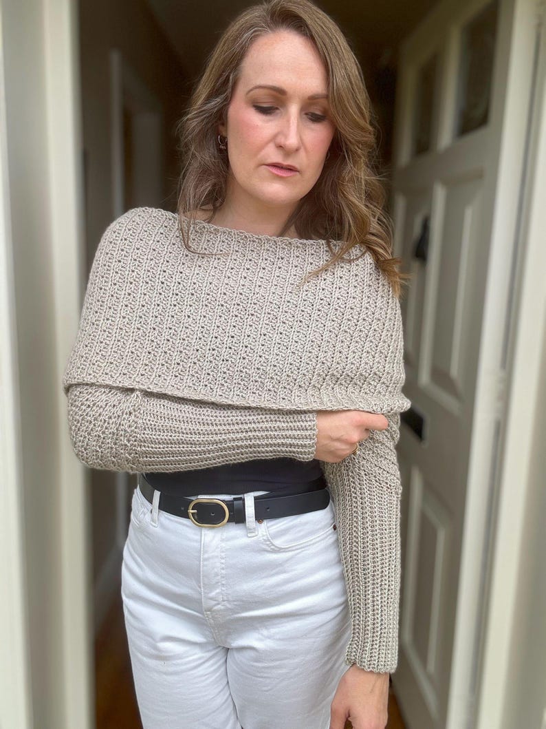 May include: A beige crochet sweater with long sleeves and a cowl neck. The sweater is worn over a black top and white jeans with a black belt.