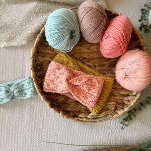 May include: A woven basket holds four balls of yarn in shades of pink, peach, and blue, along with two knitted headbands in yellow and pink. A light blue headband is also visible. The items are arranged on a white textured surface.