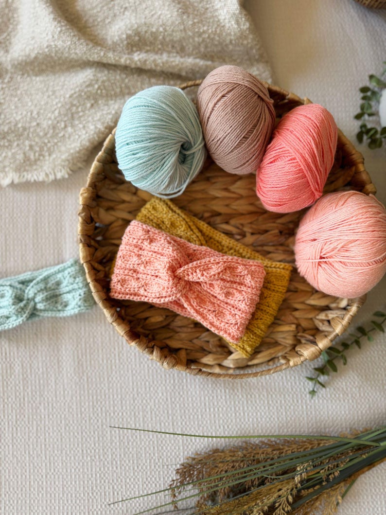 May include: A woven basket holds four balls of yarn in shades of pink, peach, and blue, along with two knitted headbands in yellow and pink. A light blue headband is also visible. The items are arranged on a white textured surface.