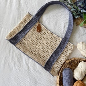 Puede incluir: Un bolso de ganchillo con cuerpo de malla calada en color blanco roto y asas s&oacute;lidas en azul pizarra. El bolso incluye una peque&ntilde;a etiqueta de aspecto cuero marr&oacute;n sujeta al borde superior. Consta de una base rectangular y dos asas largas.