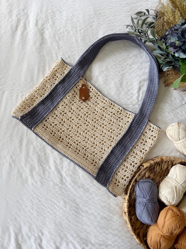 Puede incluir: Un bolso de ganchillo con cuerpo de malla calada en color blanco roto y asas s&oacute;lidas en azul pizarra. El bolso incluye una peque&ntilde;a etiqueta de aspecto cuero marr&oacute;n sujeta al borde superior. Consta de una base rectangular y dos asas largas.