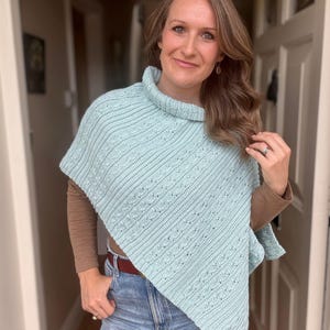 May include: A light blue knitted poncho with a high collar. The poncho features a cable knit design and is worn over a brown long-sleeved shirt and blue jeans. The poncho is a stylish and comfortable garment.