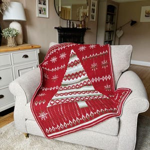 May include: A red and white crocheted throw blanket with a green and white Christmas tree design. The blanket is draped over a cream-colored armchair.