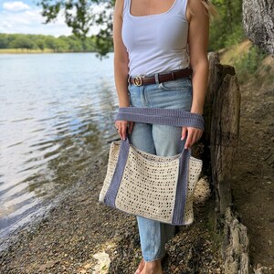 Puede incluir: Un bolso tote de ganchillo color crema con un patr&oacute;n calado repetitivo y dos asas anchas y s&oacute;lidas de color azul b&iacute;garo. El bolso tiene forma rectangular con una base estructurada y un borde azul a juego en la parte superior y los lados.