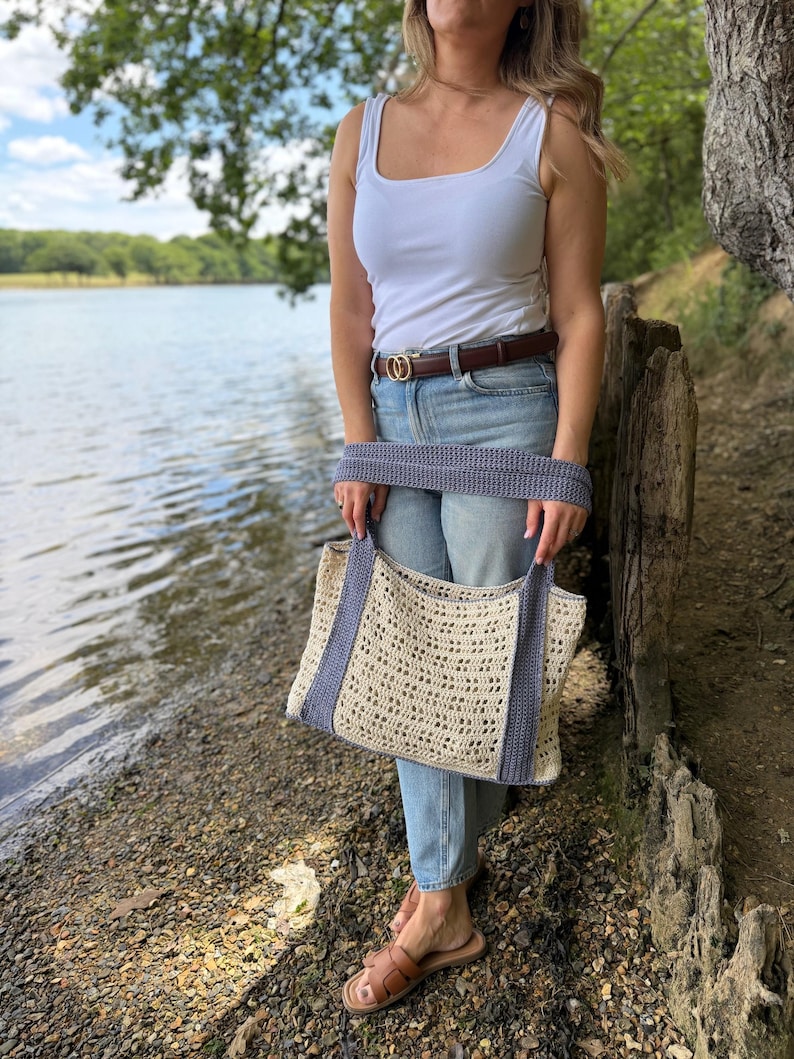 Puede incluir: Un bolso tote de ganchillo color crema con un patr&oacute;n calado repetitivo y dos asas anchas y s&oacute;lidas de color azul b&iacute;garo. El bolso tiene forma rectangular con una base estructurada y un borde azul a juego en la parte superior y los lados.