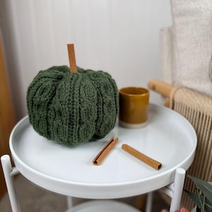May include: A green knitted pumpkin with a cinnamon stick stem sits on a white tray table. Two cinnamon sticks and a small yellow mug are also on the table. The table is part of a decorative autumn display.