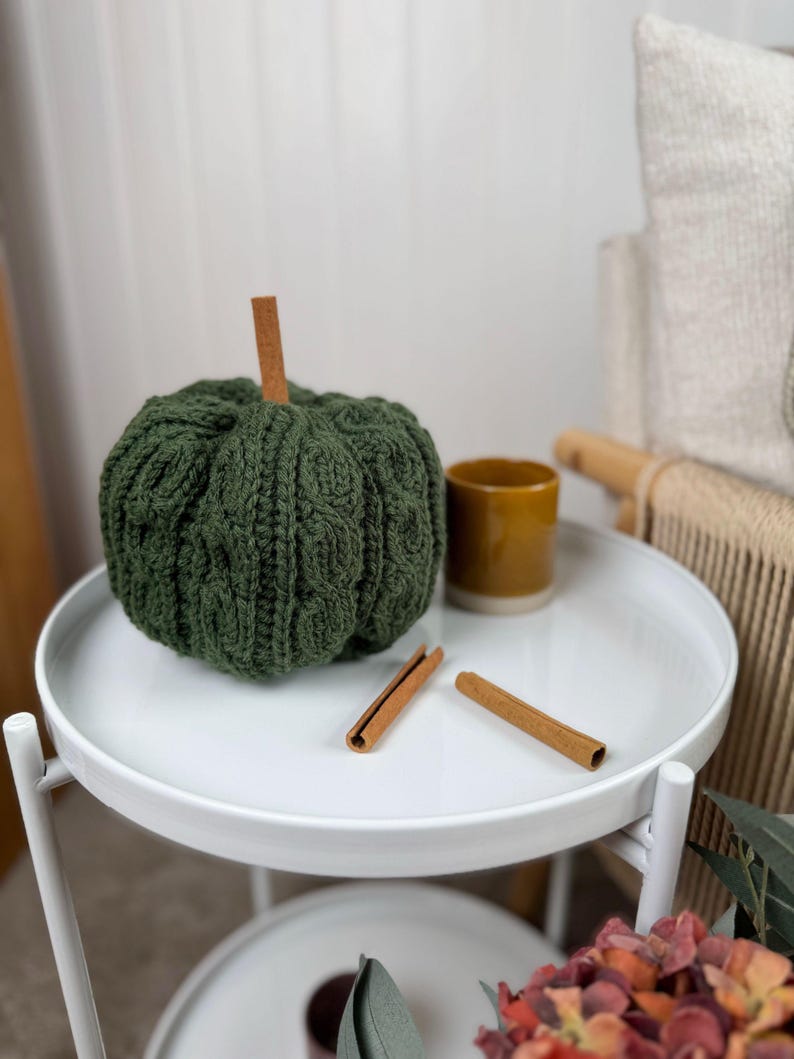 May include: A green knitted pumpkin with a cinnamon stick stem sits on a white tray table. Two cinnamon sticks and a small yellow mug are also on the table. The table is part of a decorative autumn display.