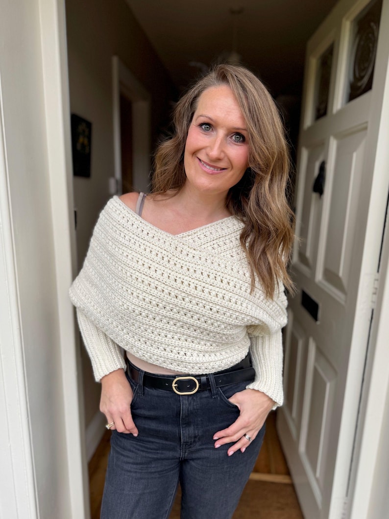 May include: A cream-colored, crocheted sweater with a V-neck and long sleeves. The sweater is paired with black jeans and a black belt with a gold oval buckle. The person is standing in front of a white door.