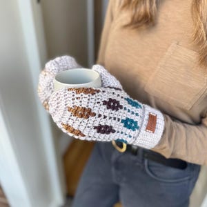 May include: Crocheted mittens in a light gray color with brown and teal geometric patterns. The mittens are holding a white ceramic mug. A small brown leather tag is sewn on the side of the mitten.