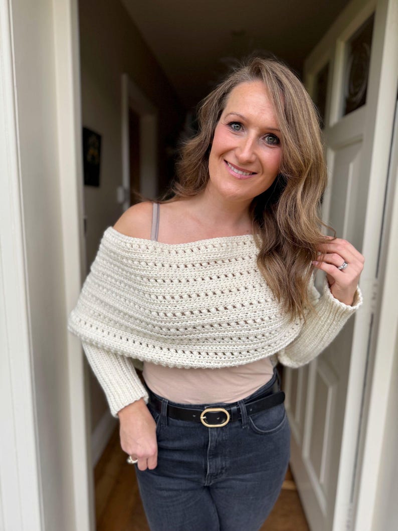 May include: Cream-colored, off-the-shoulder crochet shrug with long sleeves. The open-weave design adds texture to the garment. The model is wearing a black belt with a gold buckle and dark wash jeans. The shrug is a cozy, stylish accessory.