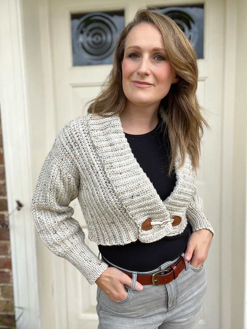 Pu&ograve; includere: Un cardigan corto con scollo a V e maniche lunghe. Il cardigan &egrave; realizzato in filato beige maculato e ha due bottoni marroni sul davanti.