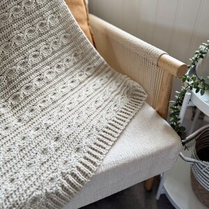 Peut inclure: Une couverture tricot&eacute;e beige clair avec un motif textur&eacute; est drap&eacute;e sur le bras d'une chaise cr&egrave;me. La couverture pr&eacute;sente un motif de tricot torsad&eacute; d&eacute;taill&eacute; et une bordure c&ocirc;tel&eacute;e. Un coussin marron repose sur la chaise.