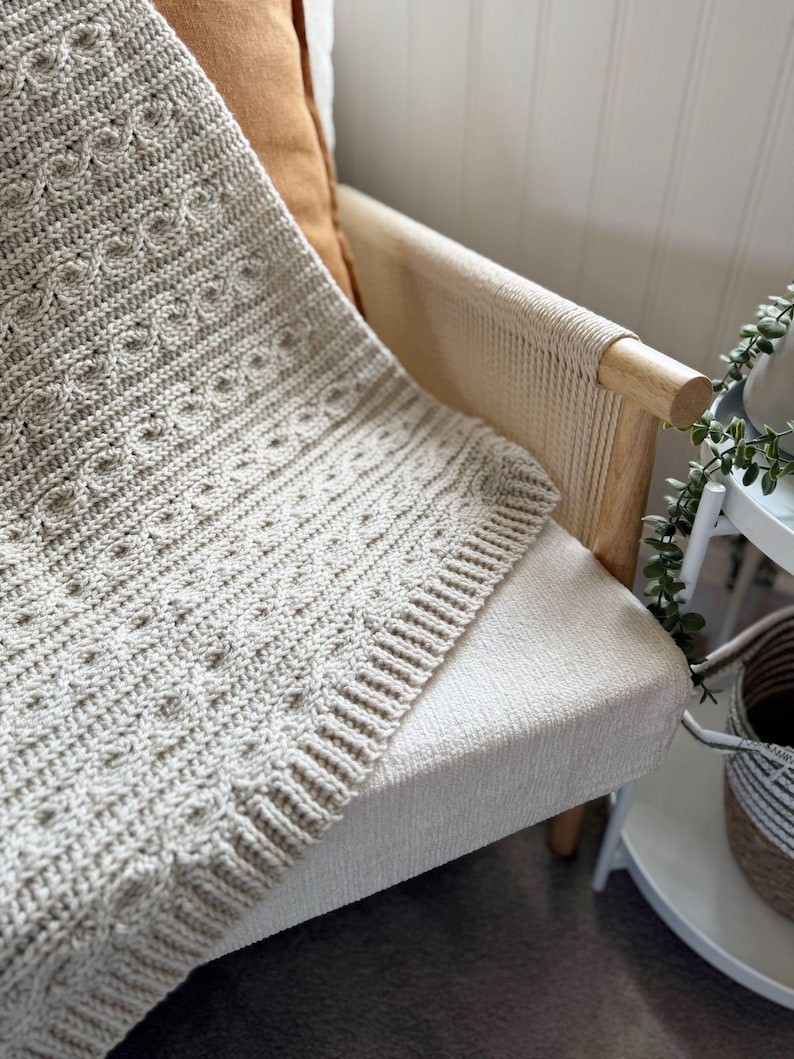 Peut inclure: Une couverture tricot&eacute;e beige clair avec un motif textur&eacute; est drap&eacute;e sur le bras d'une chaise cr&egrave;me. La couverture pr&eacute;sente un motif de tricot torsad&eacute; d&eacute;taill&eacute; et une bordure c&ocirc;tel&eacute;e. Un coussin marron repose sur la chaise.