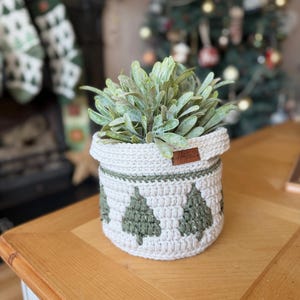 May include: A cream-coloured crochet basket featuring green Christmas tree patterns, containing a green plant. The basket is on a wooden table. A small brown tag is attached. Christmas decorations are in the background.