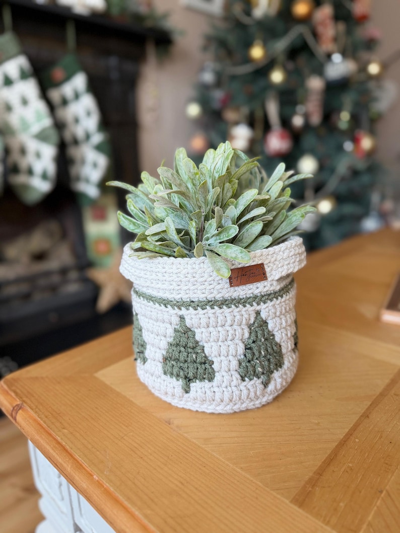 May include: A cream-coloured crochet basket featuring green Christmas tree patterns, containing a green plant. The basket is on a wooden table. A small brown tag is attached. Christmas decorations are in the background.