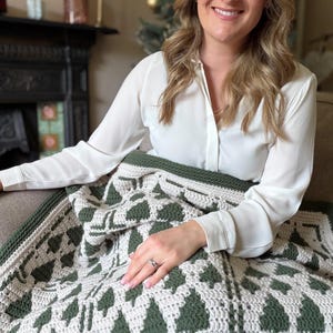 May include: A crochet blanket featuring a repeating pattern of dark forest green pine trees against a cream-colored background. The textile has a thick, textured stitch and is finished with a solid dark green border along the edges.