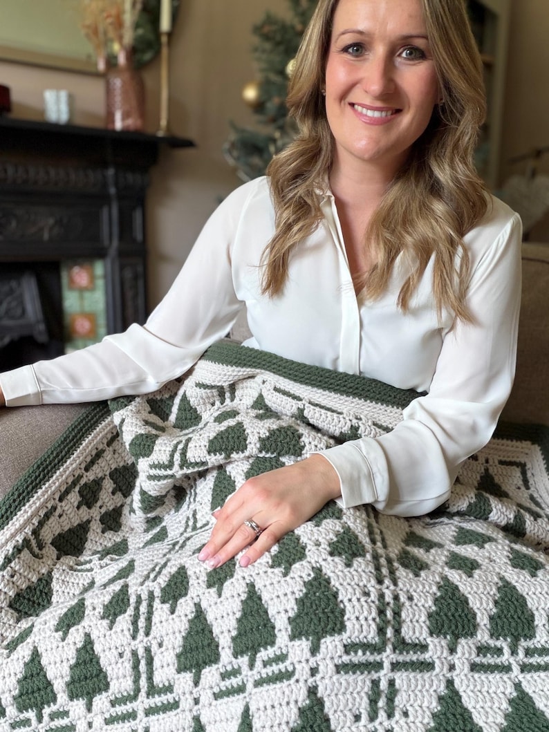 May include: A crochet blanket featuring a repeating pattern of dark forest green pine trees against a cream-colored background. The textile has a thick, textured stitch and is finished with a solid dark green border along the edges.