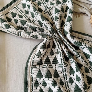 May include: A crochet blanket featuring a repeating pattern of dark forest green pine trees against a cream-colored background. The textile has a thick, textured stitch construction with a solid dark green border and a cream-colored edge trim.