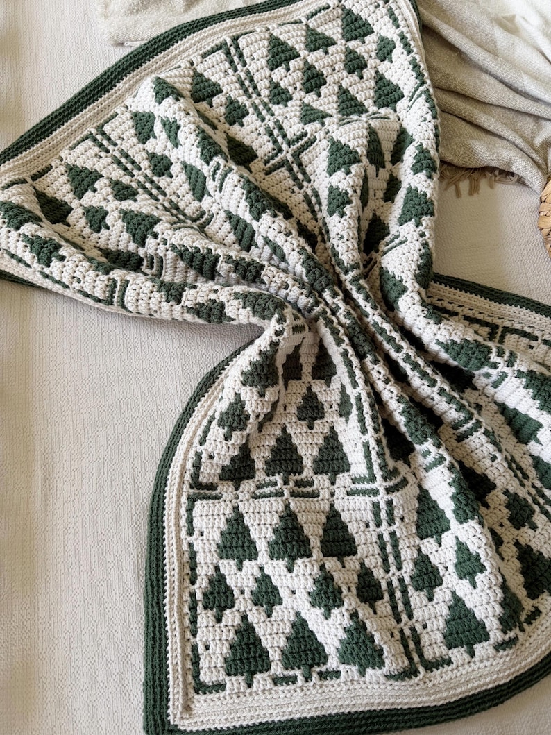 May include: A crochet blanket featuring a repeating pattern of dark forest green pine trees against a cream-colored background. The textile has a thick, textured stitch construction with a solid dark green border and a cream-colored edge trim.