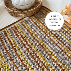 以下が含まれることがあります： 茶色、黄色、白の色合いのストライプ模様の、かぎ針編みのブランケット。ブランケットは、装飾用の白いカボチャと織りのトレイと一緒にベッドの上に展示されています。画像には「15サイズ+初心者向けビデオチュートリアル」と書かれています。
