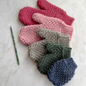 Pode incluir: Cinco pares de luvas de malha em várias cores, incluindo rosa, cinza e verde. As luvas estão dispostas em um semicírculo sobre uma superfície branca.