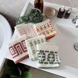 May include: Six handmade crochet washcloths in cream, pink, orange, burgundy, and green, each with a unique geometric pattern. The washcloths are displayed on a white sink, showcasing their intricate designs.
