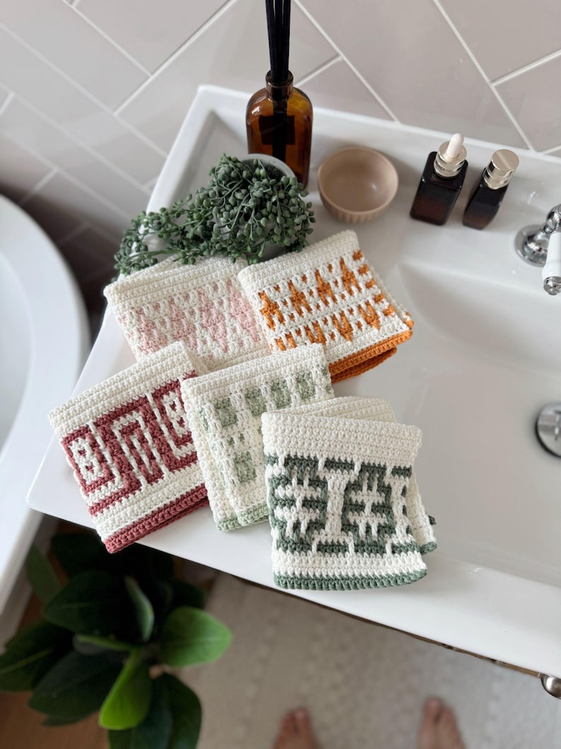 May include: Six handmade crochet washcloths in cream, pink, orange, burgundy, and green, each with a unique geometric pattern. The washcloths are displayed on a white sink, showcasing their intricate designs.