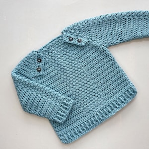 May include: A light blue crocheted sweater with a button closure at the neck. The sweater has long sleeves and a ribbed hem.