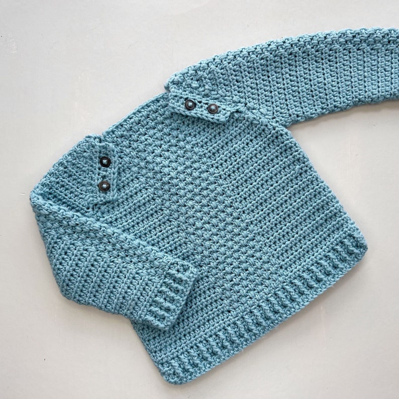 May include: A light blue crocheted sweater with a button closure at the neck. The sweater has long sleeves and a ribbed hem.