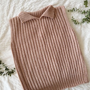 May include: A pale brown, collared, sleeveless top. The top is made of a ribbed knit material. The collar is a classic polo style. The garment is displayed on a white surface with green foliage in the background.