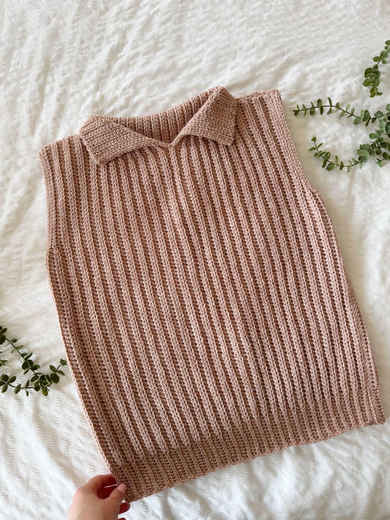 May include: A pale brown, collared, sleeveless top. The top is made of a ribbed knit material. The collar is a classic polo style. The garment is displayed on a white surface with green foliage in the background.