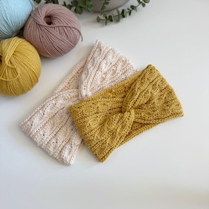 May include: Three knitted headbands in cream, mustard yellow, and light beige, with a cable knit design. The headbands are displayed on a white surface, with balls of yarn in pastel colors in the background. The headbands are a stylish accessory.