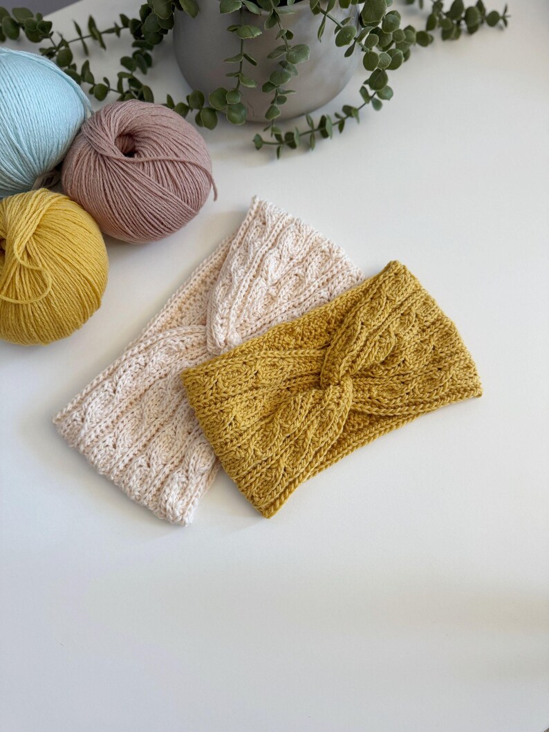 May include: Three knitted headbands in cream, mustard yellow, and light beige, with a cable knit design. The headbands are displayed on a white surface, with balls of yarn in pastel colors in the background. The headbands are a stylish accessory.