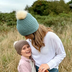 May include: A light teal knitted beanie with a cream-colored faux fur pom-pom. A child wears a brown knitted beanie with a cream-colored faux fur pom-pom. The child is wearing a pink button-down shirt.