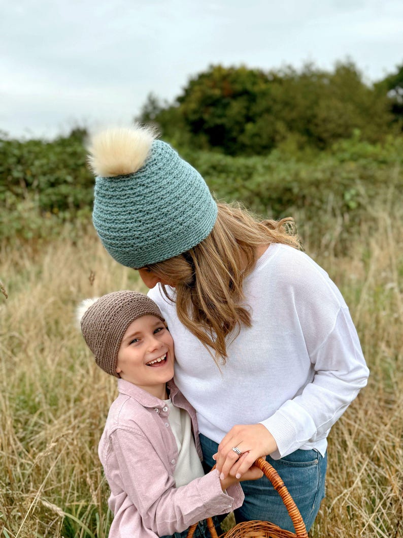 May include: A light teal knitted beanie with a cream-colored faux fur pom-pom. A child wears a brown knitted beanie with a cream-colored faux fur pom-pom. The child is wearing a pink button-down shirt.