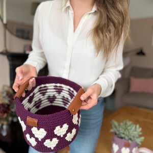 Op de afbeelding: Een handgemaakte gehaakte mand in diep paars met witte hartpatronen. De mand heeft bruine leren handvatten en een klein label. Extra gehaakte plantenbakken met hartontwerpen zijn zichtbaar op de achtergrond.