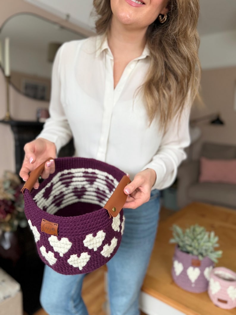 Op de afbeelding: Een handgemaakte gehaakte mand in diep paars met witte hartpatronen. De mand heeft bruine leren handvatten en een klein label. Extra gehaakte plantenbakken met hartontwerpen zijn zichtbaar op de achtergrond.