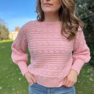 Puede incluir: Un suéter de color rosa tejido a crochet con un corte holgado y mangas largas. El suéter tiene un delicado patrón de puntadas y un cuello ligeramente de barco.