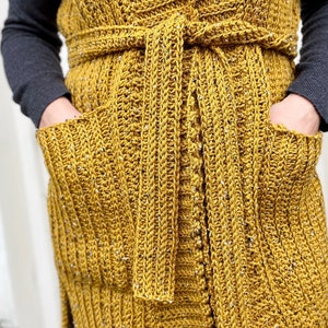 Pu&ograve; includere: Un lungo gilet di uncinetto giallo senape con una cintura a cravatta e due tasche. Il gilet ha un motivo testurizzato e a coste.