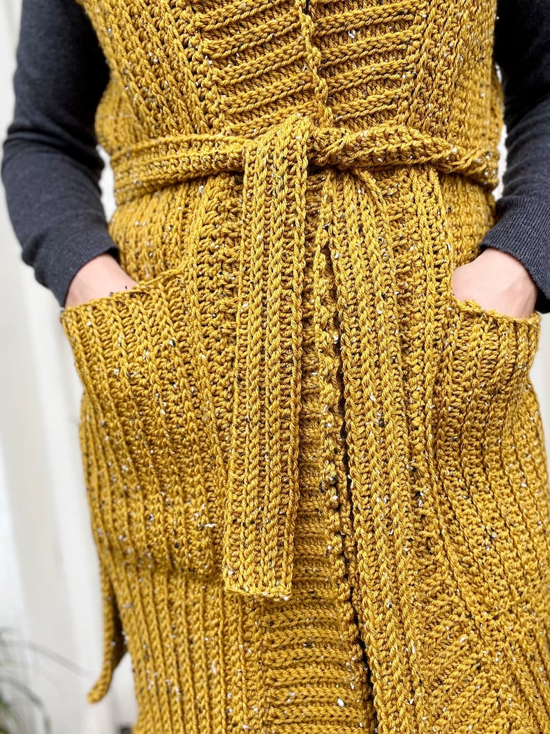 Pu&ograve; includere: Un lungo gilet di uncinetto giallo senape con una cintura a cravatta e due tasche. Il gilet ha un motivo testurizzato e a coste.