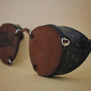 May include: Round eyewear featuring dark brown wooden lenses and black side shields. The side shields are constructed with a series of small metal eyelets. The wooden lenses are held in place by thin silver-toned metal wire frames and connectors.
