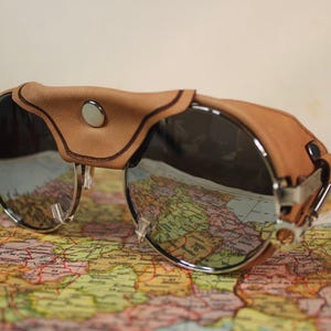 May include: Round sunglasses featuring dark tinted lenses and a silver metal frame. The eyewear includes tan leather side shields and a matching leather bridge cover secured with a central silver snap button, providing a distinct look.